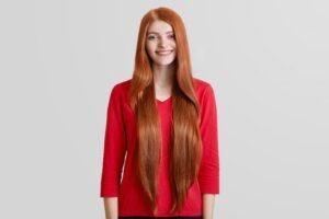 cheerful-beautiful-female-with-very-long-red-hair-freckled-face-dressed-casual-red-sweater-poses-against-white-wall-has-pleasant-gentle-smile-positivity-beauty-style-concept_176532-6519